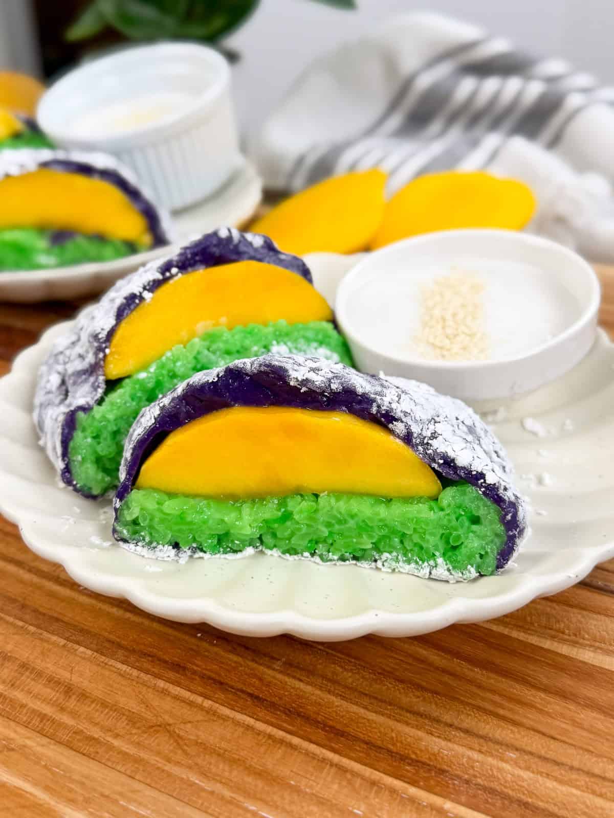 Sliced mango, green sticky rice, and purple rice wrapped in silver foil on a plate, with a small bowl of white sauce topped with sesame seeds—this vibrant presentation puts a unique spin on Mango Sticky Rice Ube Mochi.