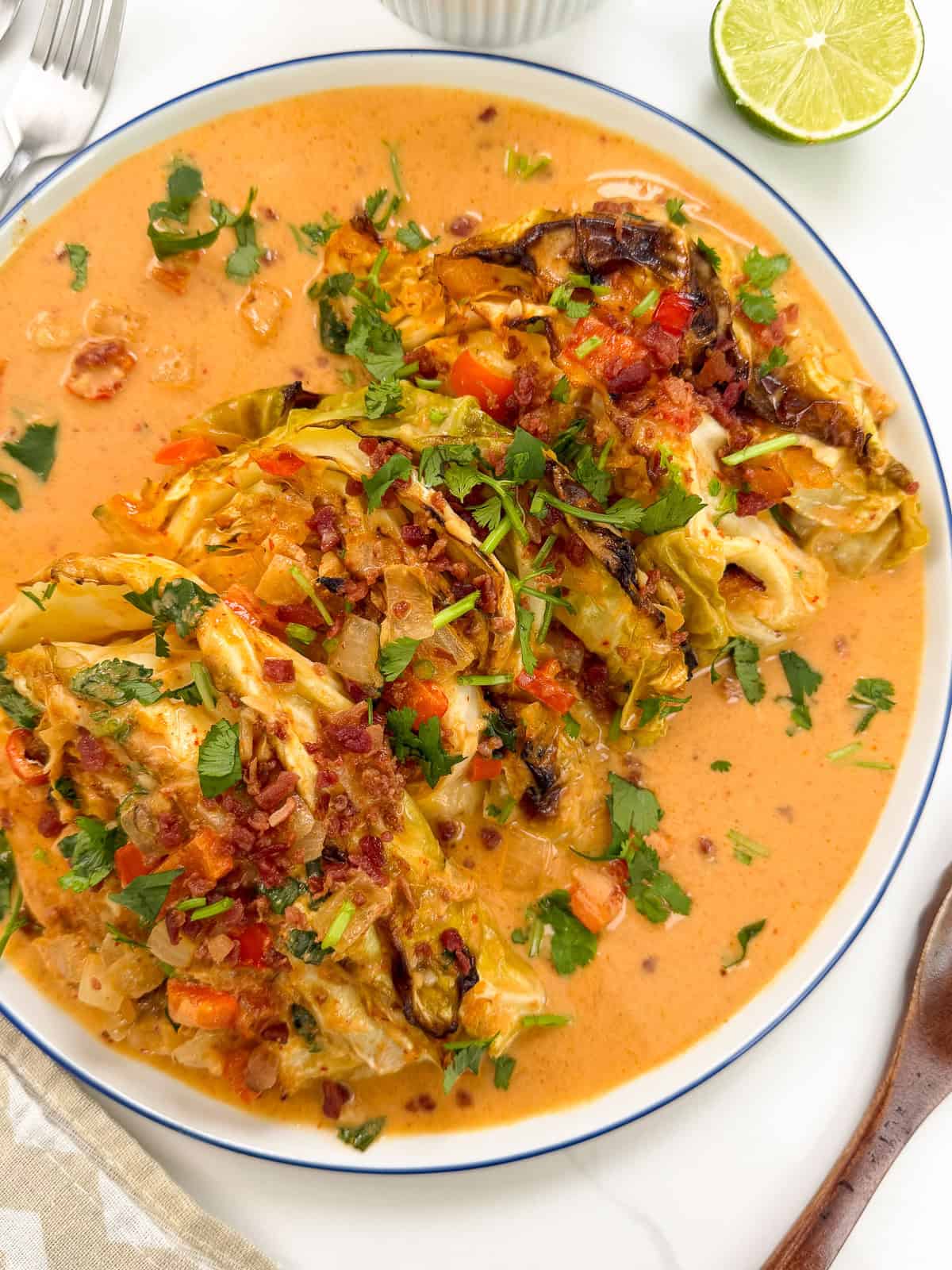 A plate of Red Curry Melting Cabbage is presented in a creamy, orange-colored sauce, garnished with chopped herbs and crispy bits, with a lime half nearby on the table.