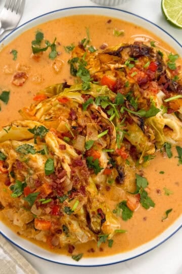 A plate of Red Curry Melting Cabbage wedges topped with chopped vegetables and herbs, served in a creamy orange-colored sauce. Sliced lime, cutlery, and a napkin are visible nearby.