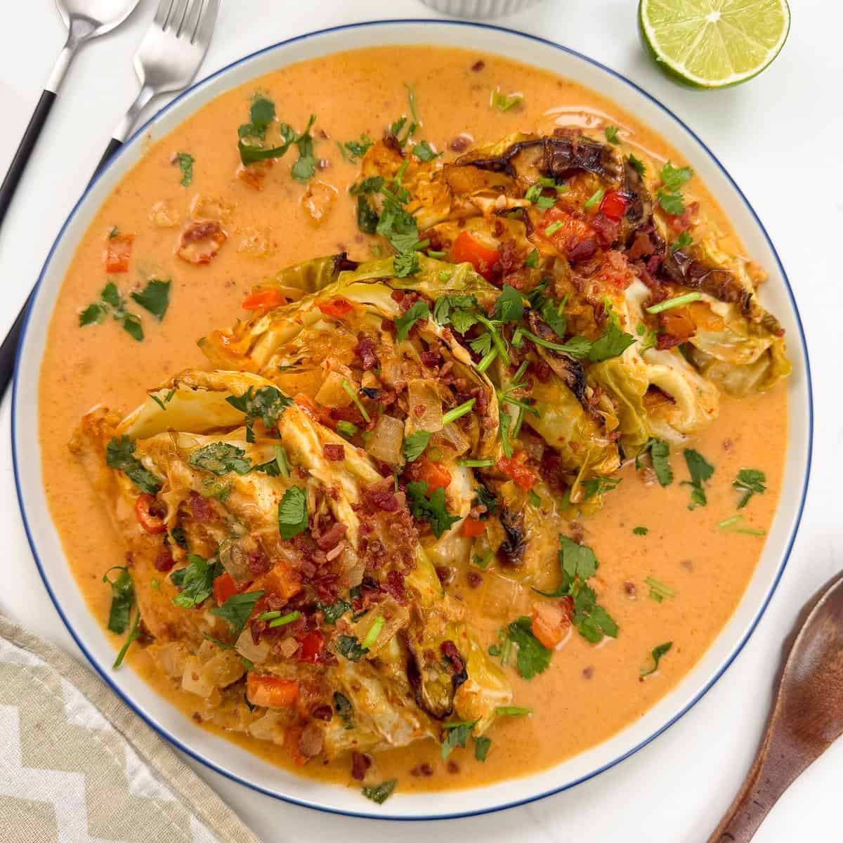 A plate of Red Curry Melting Cabbage wedges topped with chopped vegetables and herbs, served in a creamy orange-colored sauce. Sliced lime, cutlery, and a napkin are visible nearby.
