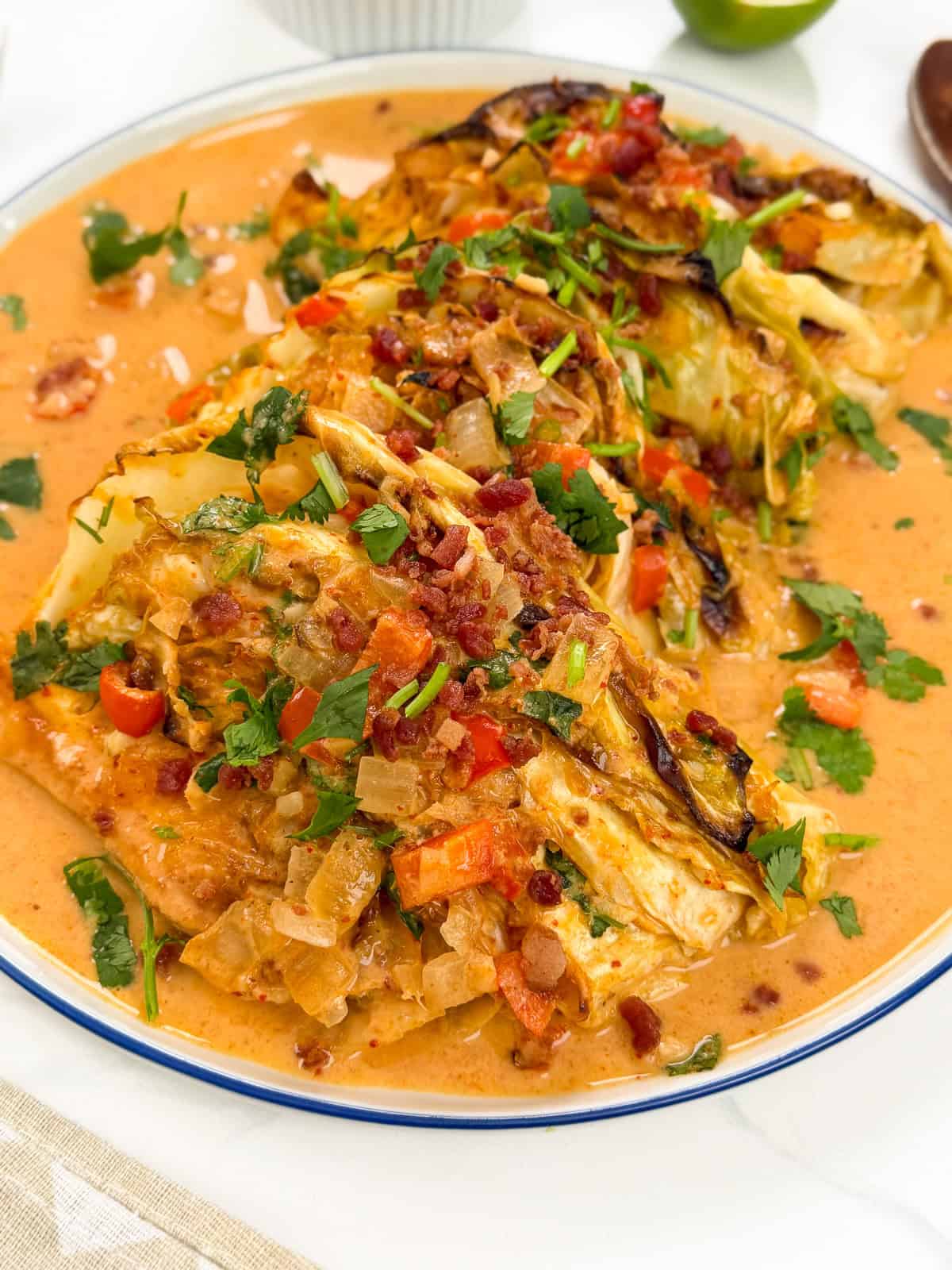 Red Curry Melting Cabbage wedges are roasted and topped with chopped vegetables, fresh herbs, and crumbled bacon, then served in a creamy orange sauce on a white plate.