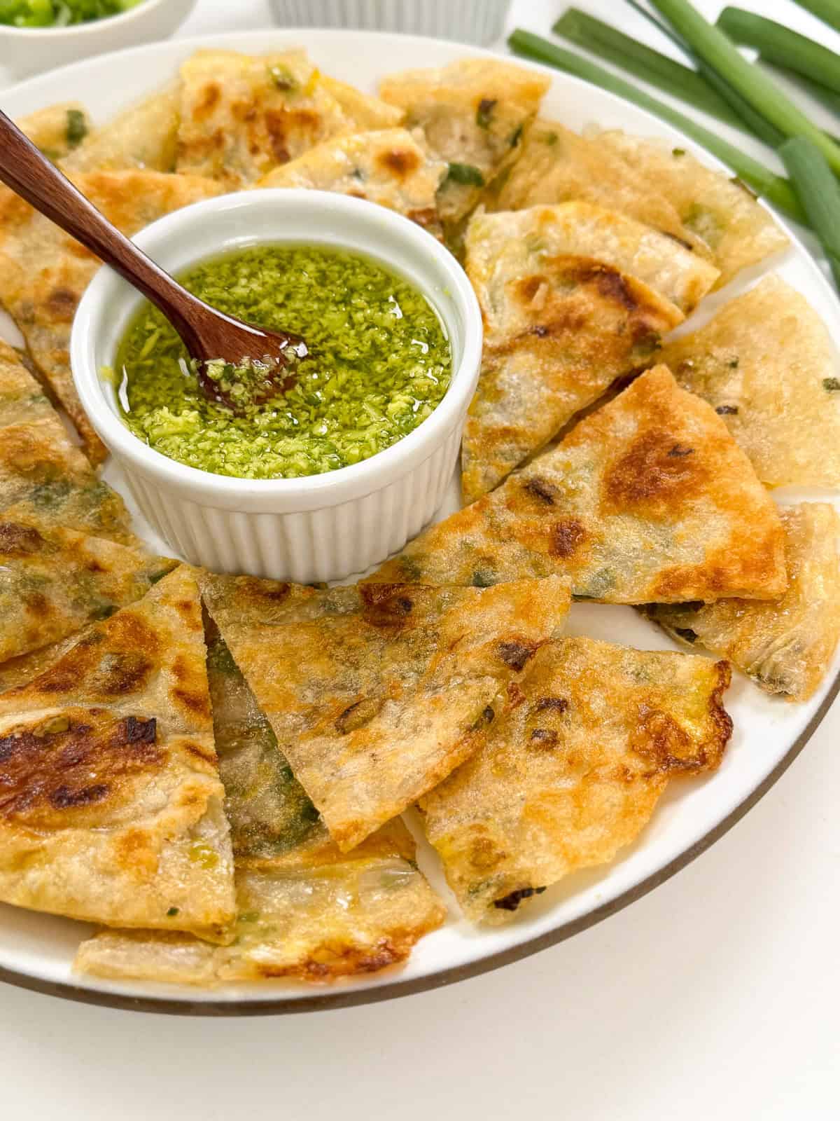 A plate of golden-brown Rice Paper Scallion Pancakes, sliced and arranged in a circle around a small bowl of green dipping sauce with a wooden spoon. Fresh green onions can be seen in the background.