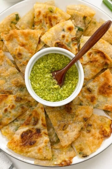 A plate of sliced, golden-brown Rice Paper Scallion Pancakes arranged around a small bowl of green dipping sauce, with a wooden spoon resting in the sauce. Green onions and chopsticks are nearby.