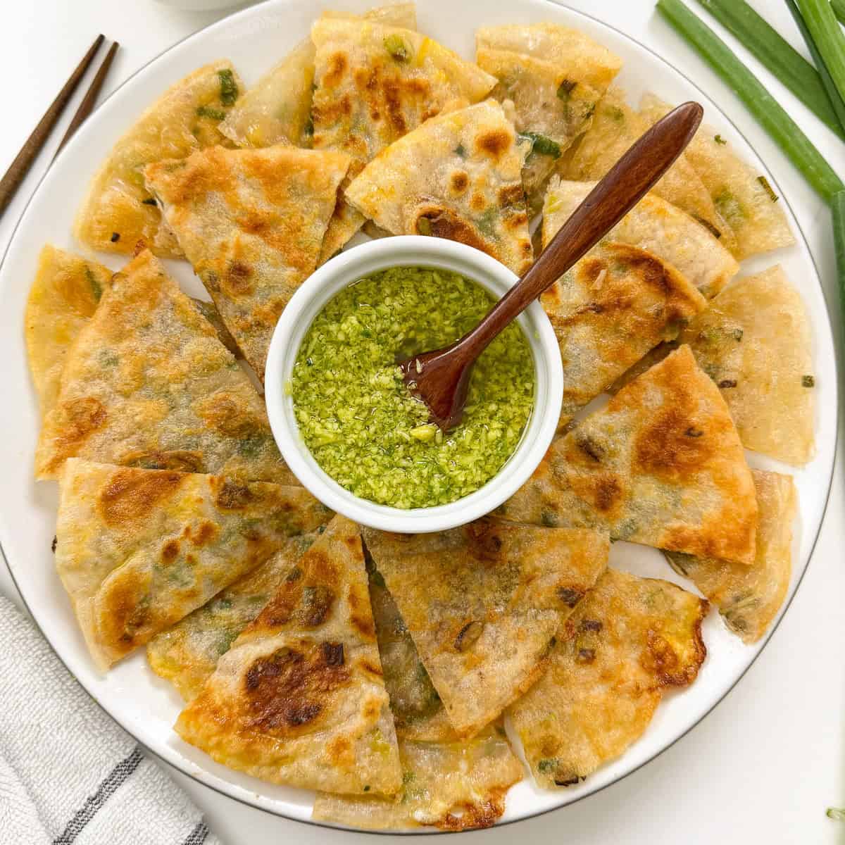 A plate of sliced, golden-brown Rice Paper Scallion Pancakes arranged around a small bowl of green dipping sauce, with a wooden spoon resting in the sauce. Green onions and chopsticks are nearby.
