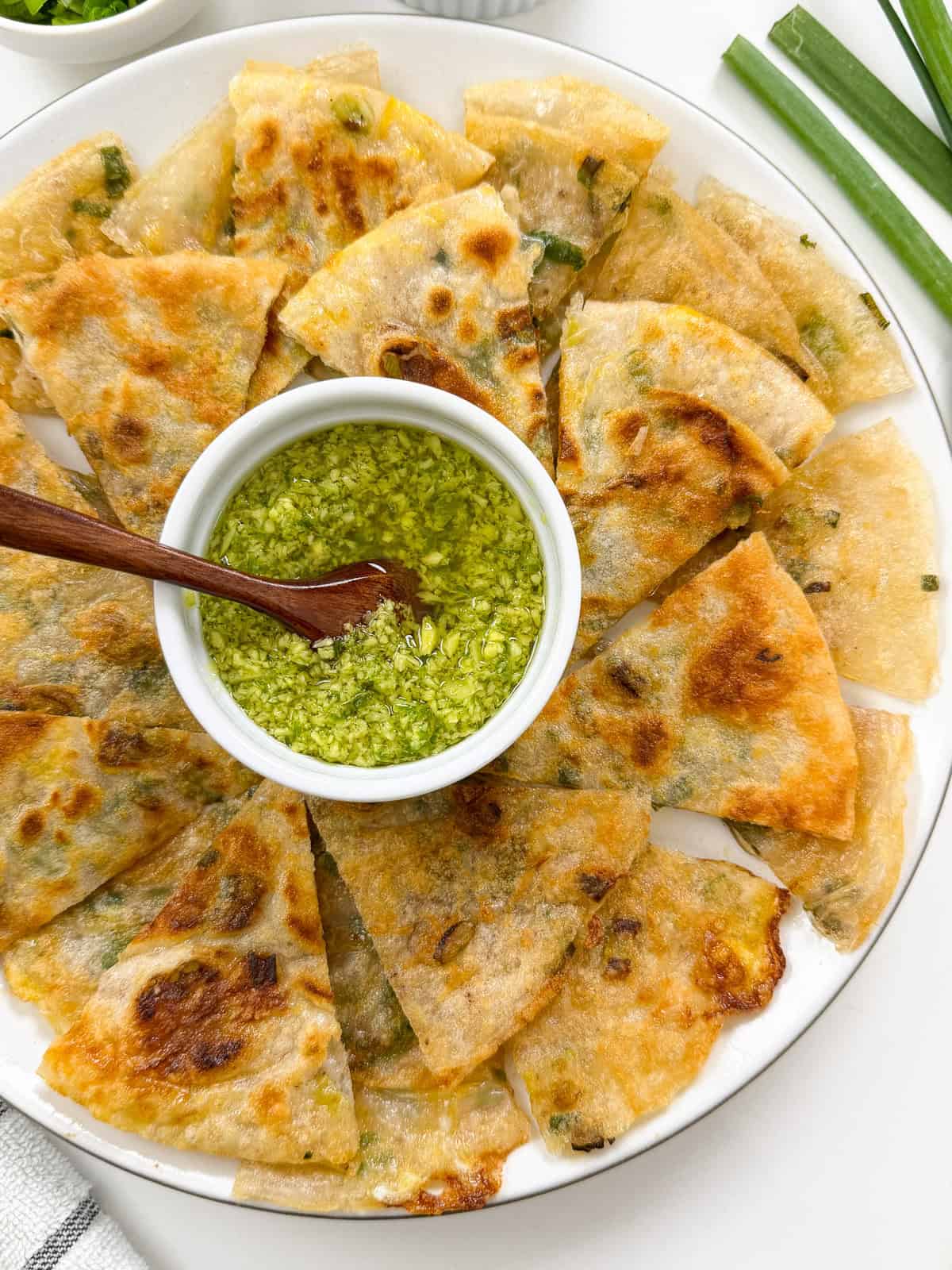 A plate of golden, crispy rice paper scallion pancakes cut into triangles, arranged around a small bowl of green dipping sauce with a wooden spoon. Green onions are visible as garnish on the side.