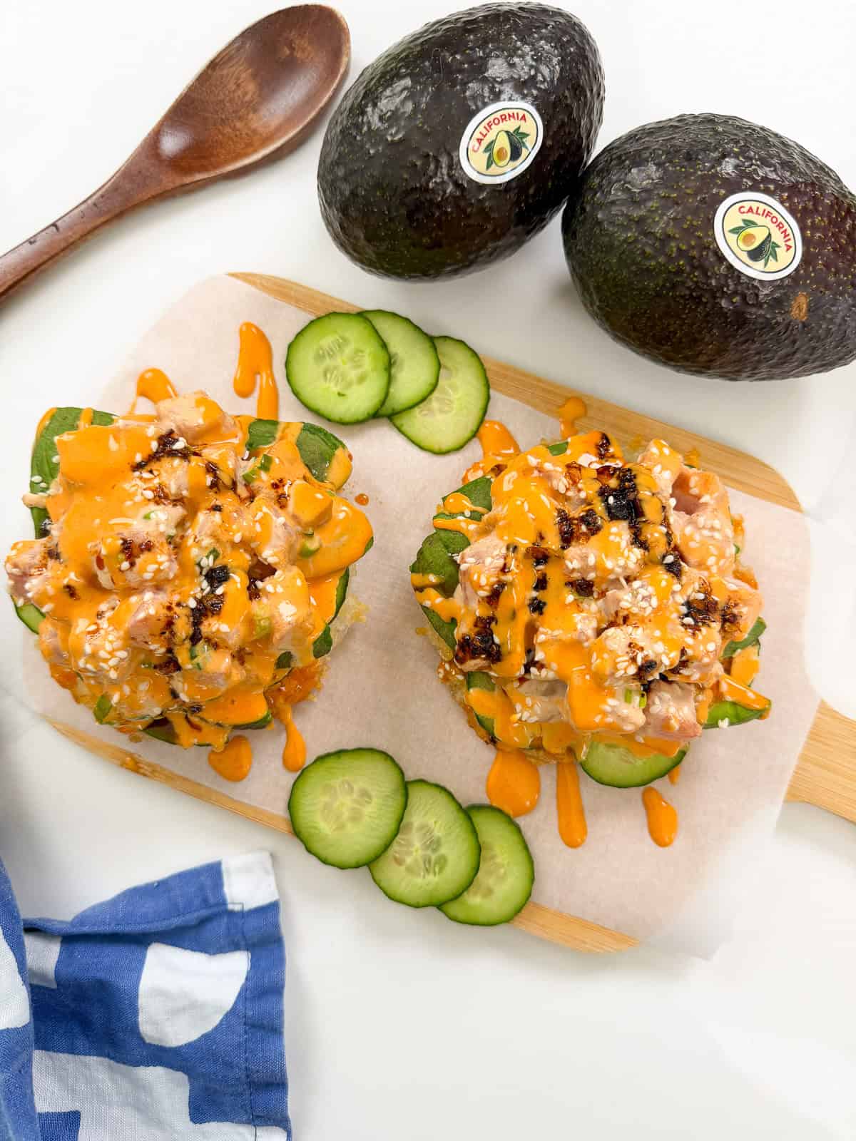Two servings of spicy tuna poke on a wooden board with sliced cucumbers, two whole avocados, and a wooden spoon nearby. Inspired by the vibrant fusion of Sushi Waffles, the poke is topped with orange sauce and sesame seeds.
