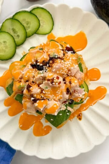 A plate with diced sushi-grade fish, avocado, and cucumber, topped with orange sauce, sesame seeds, and chili oil, served atop crispy sushi waffles with cucumber slices on the side.