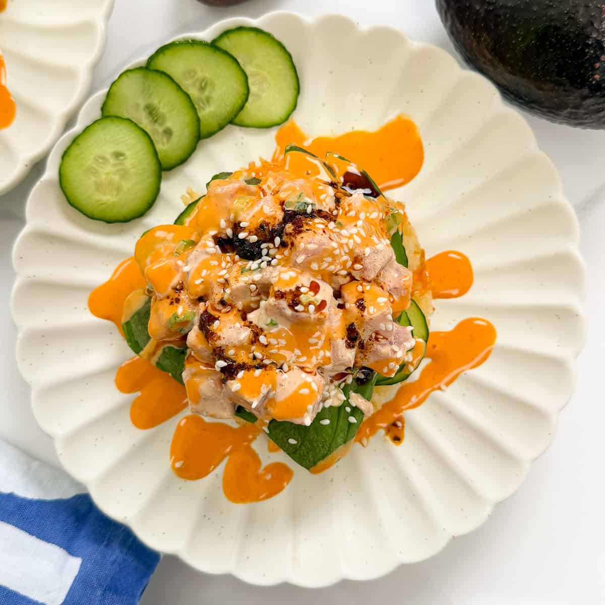 A plate with diced sushi-grade fish, avocado, and cucumber, topped with orange sauce, sesame seeds, and chili oil, served atop crispy sushi waffles with cucumber slices on the side.