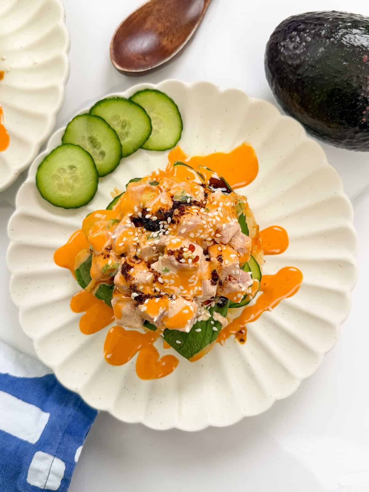 A white scalloped plate with diced fish, avocado, cucumber slices, drizzled orange sauce, sesame seeds, and chili flakes—reminiscent of the vibrant toppings on sushi waffles. A wooden spoon, avocado, and blue-white napkin are nearby.