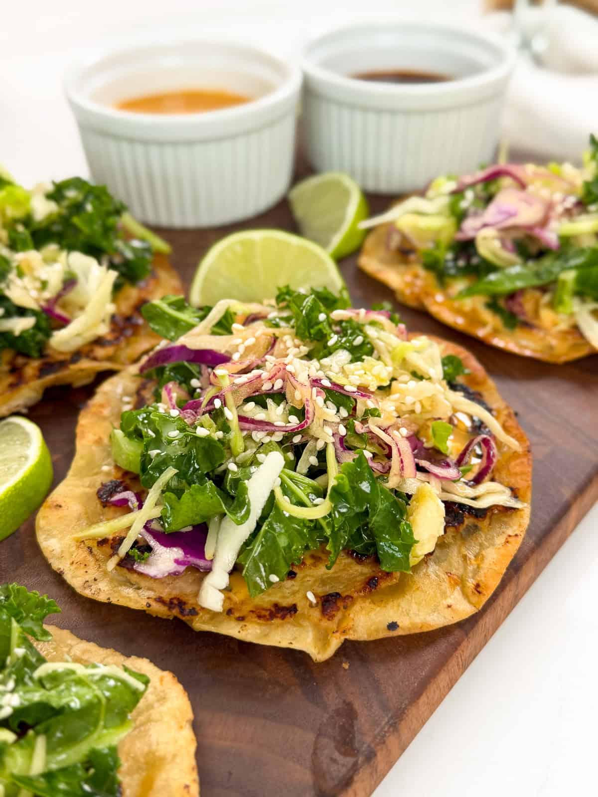 Tostadas topped with mixed greens, shredded cabbage, and pickled onions on a wooden board, inspired by the bold flavors of Sweet Chili Chicken Smashed Tacos, with lime wedges and two small cups of dipping sauces in the background.