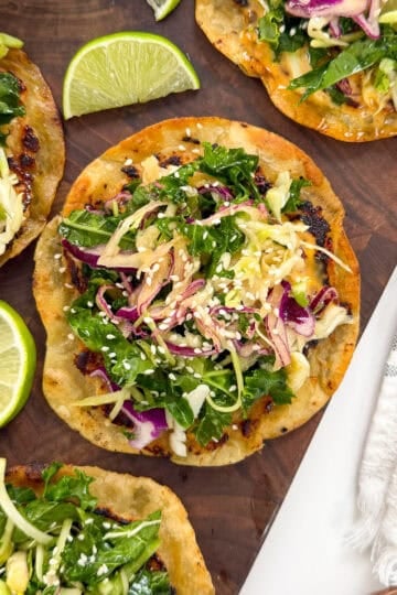 Crispy tostadas topped with greens, shredded vegetables, and sesame seeds are arranged on a wooden board, garnished with lime wedges. Inspired by the flavors of Sweet Chili Chicken Smashed Tacos, this vibrant dish is perfect for sharing. A striped napkin is beside the board.