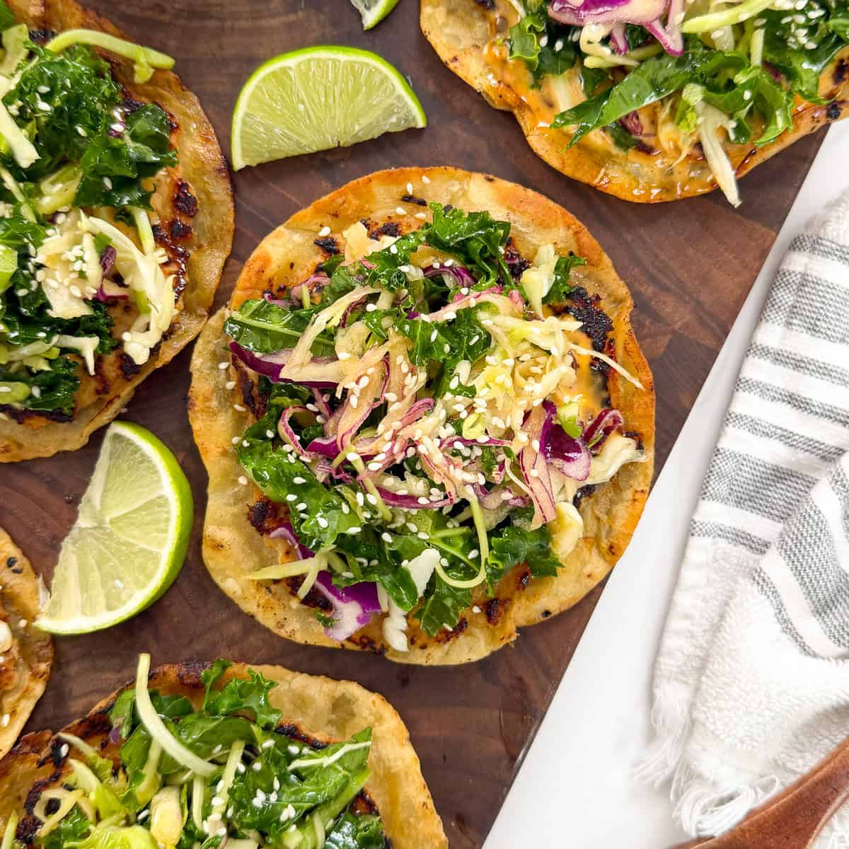 Crispy tostadas topped with greens, shredded vegetables, and sesame seeds are arranged on a wooden board, garnished with lime wedges. Inspired by the flavors of Sweet Chili Chicken Smashed Tacos, this vibrant dish is perfect for sharing. A striped napkin is beside the board.