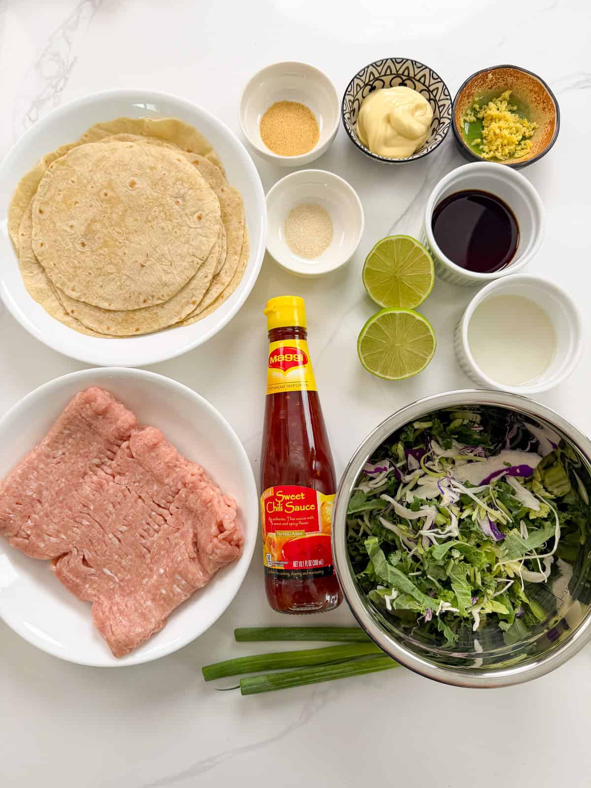 Ingredients for Sweet Chili Chicken Smashed Tacos are arranged on a white surface: tortillas, ground meat, sweet chili sauce, shredded greens, green onion, lime halves, mayo, garlic, seasonings, soy sauce, and a small bowl of rice vinegar.
