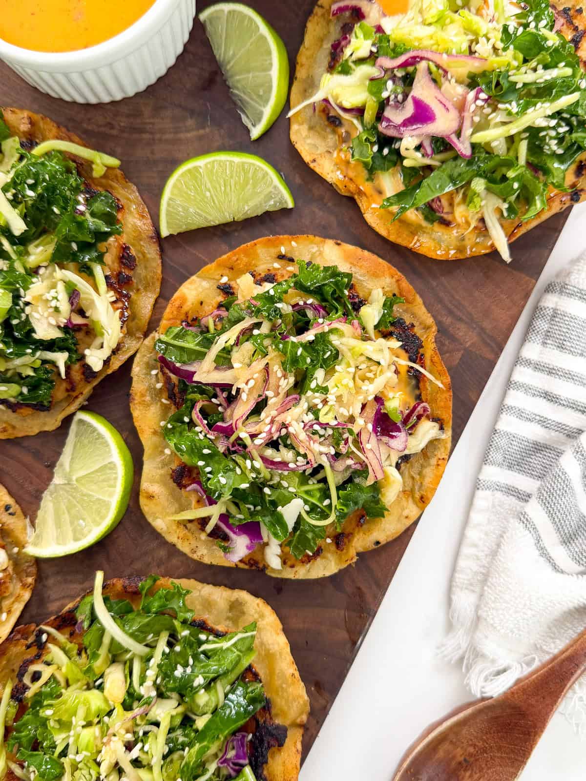 Tostadas topped with shredded greens, cabbage, cheese, and sesame seeds are arranged on a wooden board with lime wedges and a small bowl of orange sauce on the side—perfect alongside Sweet Chili Chicken Smashed Tacos for a colorful and flavorful spread. A striped napkin is at the edge.