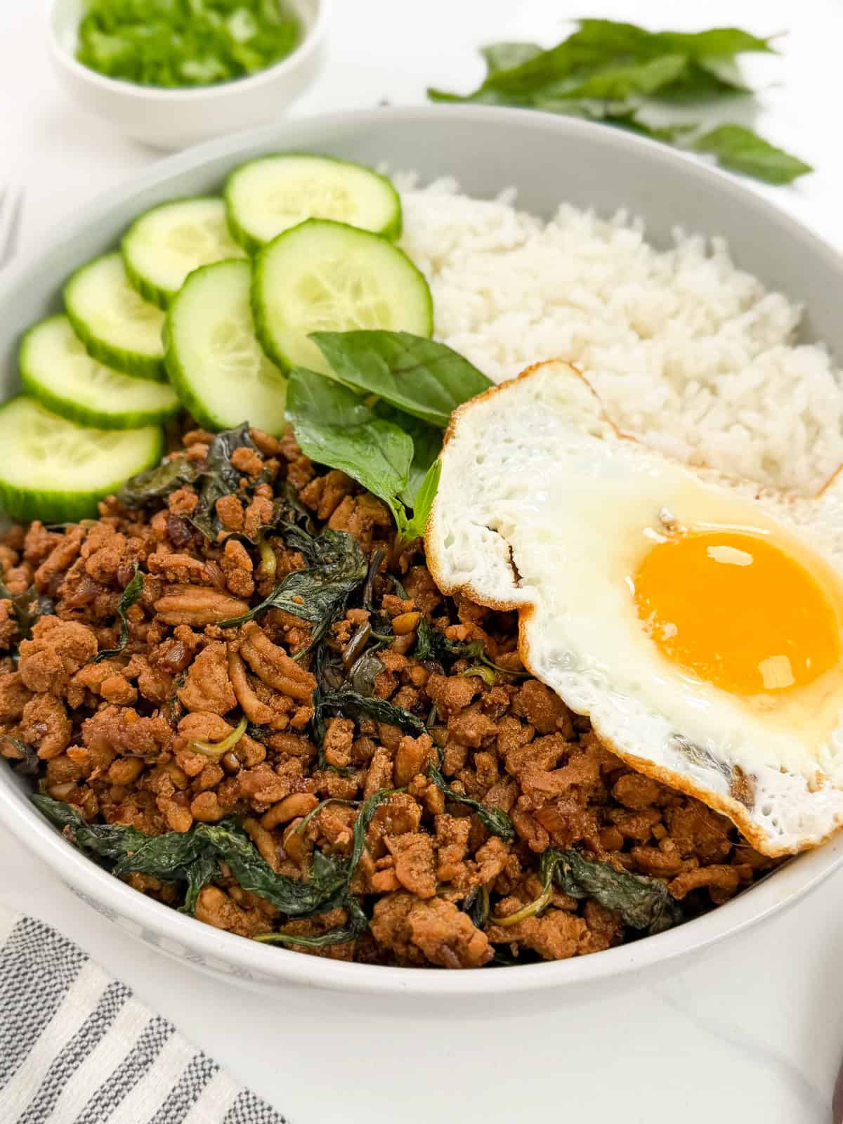 A bowl of white rice, flavorful Thai Basil Chicken stir-fried with minced meat and basil, sliced cucumber, and a sunny side up egg, garnished with fresh herbs.