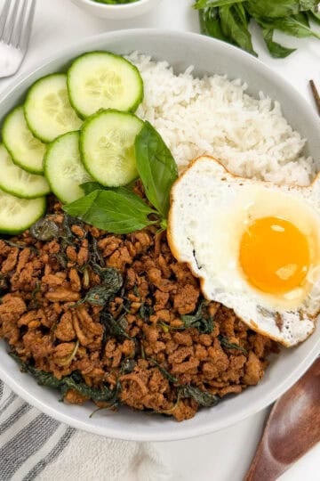 A bowl with white rice, Thai Basil Chicken stir-fried with herbs, cucumber slices, and a sunny-side-up egg. Chopsticks, a spoon, and fresh herbs are nearby.