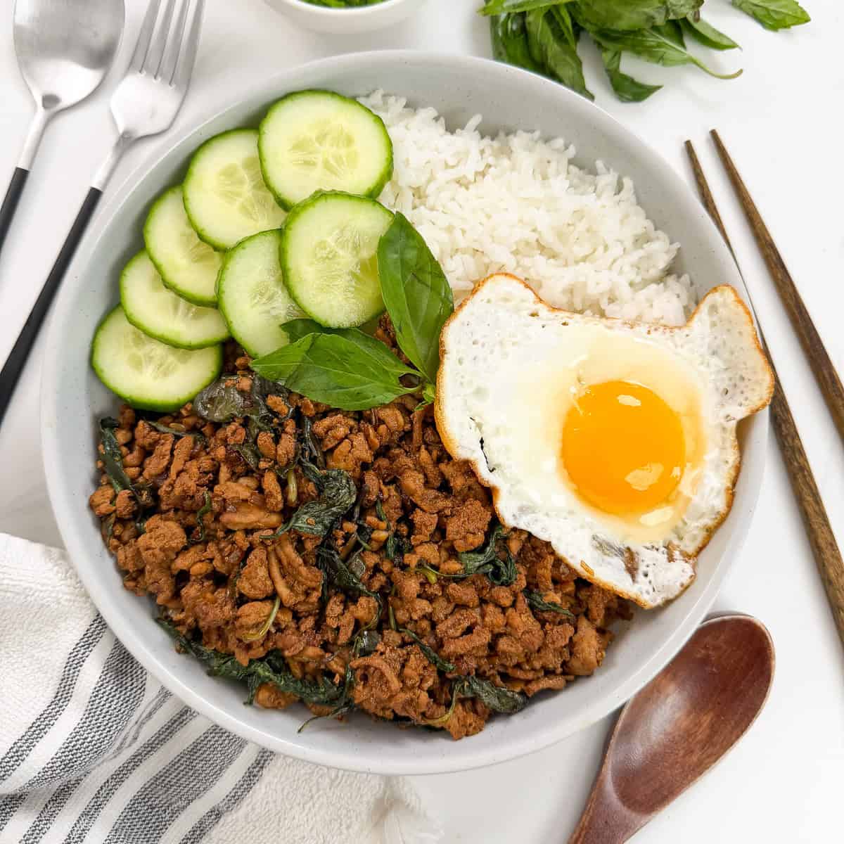 A bowl with white rice, Thai Basil Chicken stir-fried with herbs, cucumber slices, and a sunny-side-up egg. Chopsticks, a spoon, and fresh herbs are nearby.