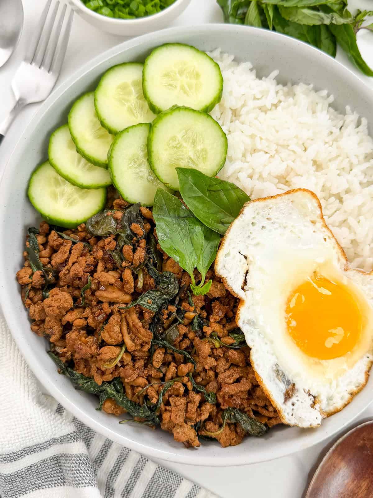 A bowl with white rice, savory Thai Basil Chicken ground meat cooked with basil, fresh cucumber slices, and a sunny-side-up fried egg on top.