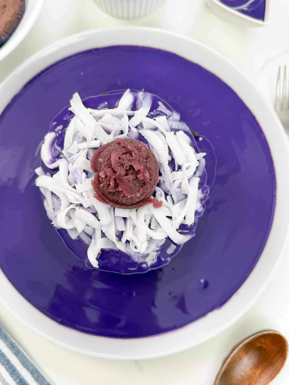 A dessert served on a purple plate, featuring shaved ice topped with shredded coconut and a scoop of dark purple ice cream, reminiscent of Ube Pancakes with Ube Coconut Sauce, with utensils and bowls visible around the plate.