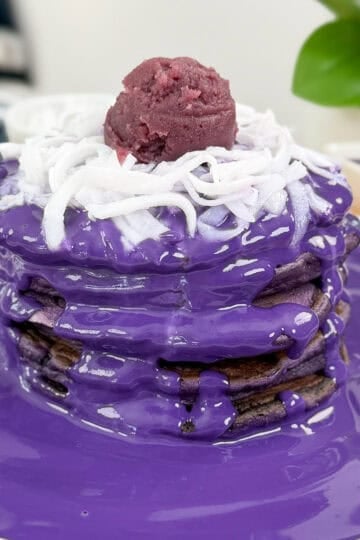 A stack of Ube Pancakes with Ube Coconut Sauce, topped with shredded coconut and a scoop of purple yam paste, sits on a white plate with extra sauce pooled around. A green plant decorates the background.