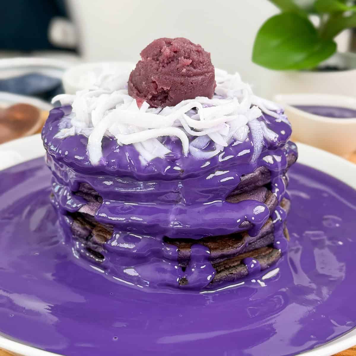 A stack of Ube Pancakes with Ube Coconut Sauce, topped with shredded coconut and a scoop of purple yam paste, sits on a white plate with extra sauce pooled around. A green plant decorates the background.