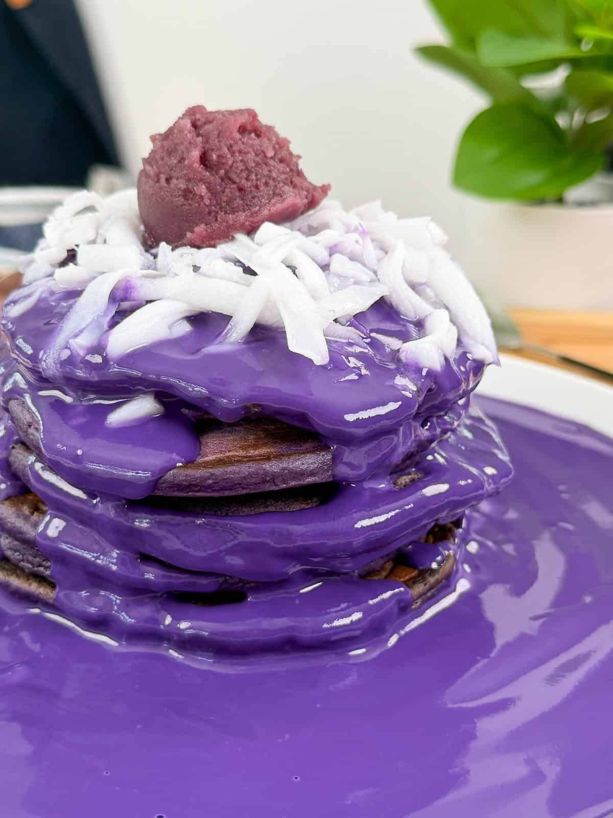 A stack of ube pancakes with ube coconut sauce, covered in glossy purple sauce, topped with shredded coconut and a scoop of purple paste, sits on a white plate with a green plant in the background.
