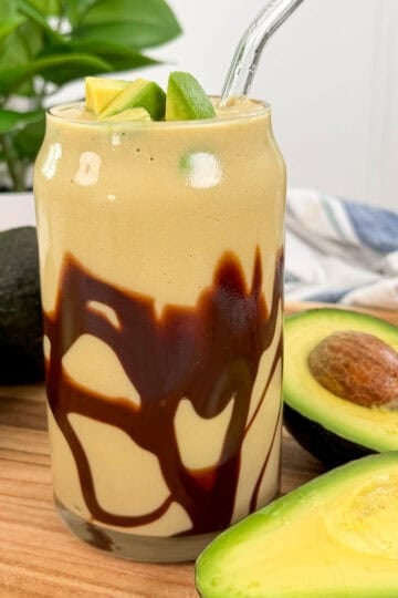 A creamy Avocado Frappe (Blended Ice Coffee) smoothie in a glass with chocolate syrup drizzled inside, topped with avocado slices and a clear straw. Whole and halved avocados are nearby on a wooden surface.