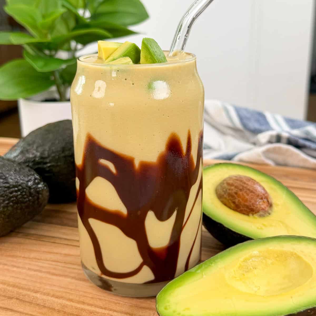 A creamy Avocado Frappe (Blended Ice Coffee) smoothie in a glass with chocolate syrup drizzled inside, topped with avocado slices and a clear straw. Whole and halved avocados are nearby on a wooden surface.