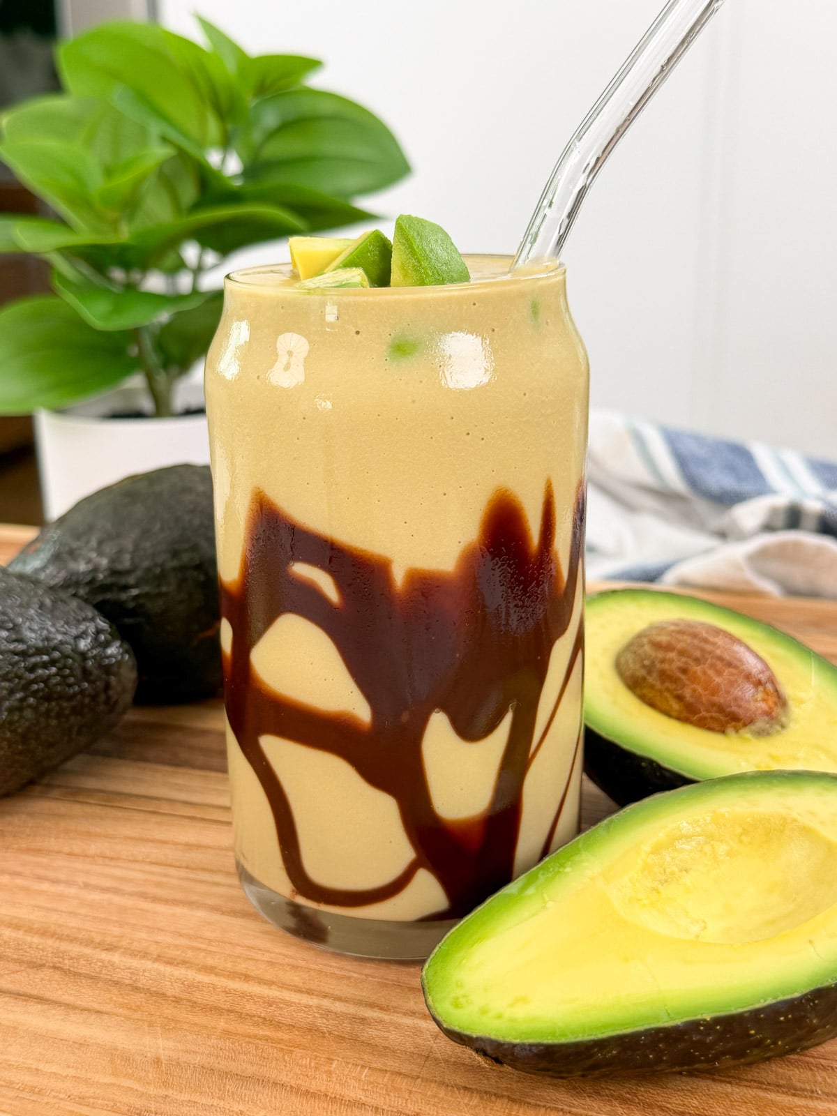 A glass filled with a creamy Avocado Coffee Smoothie, drizzled with chocolate syrup, garnished with avocado chunks, and a glass straw. Fresh avocados and a green plant are in the background on a wooden surface.