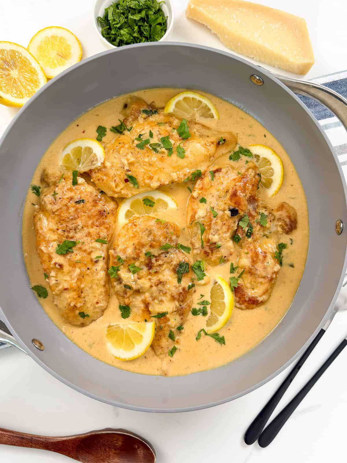 Chicken breasts simmer in a creamy sauce with lemon slices and chopped herbs in a gray skillet, creating an inviting Creamy Lemon Garlic Chicken. Fresh parsley, lemon, and parmesan are nearby. The scene is bright and appetizing with utensils on the side.