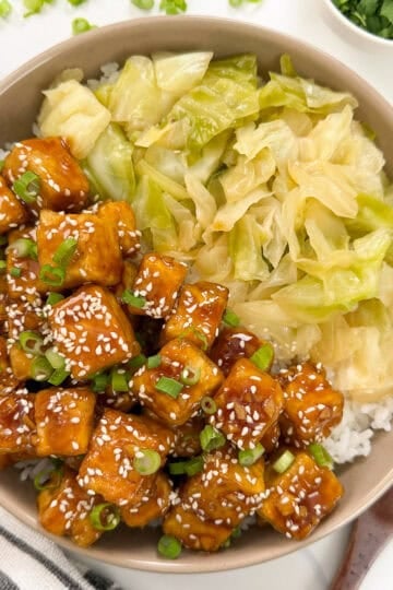 A bowl of rice topped with Honey Butter Crispy Tofu cubes, glazed and sprinkled with sesame seeds and green onions, sits alongside sautéed cabbage. Chopsticks, a fork, and a napkin are nearby.
