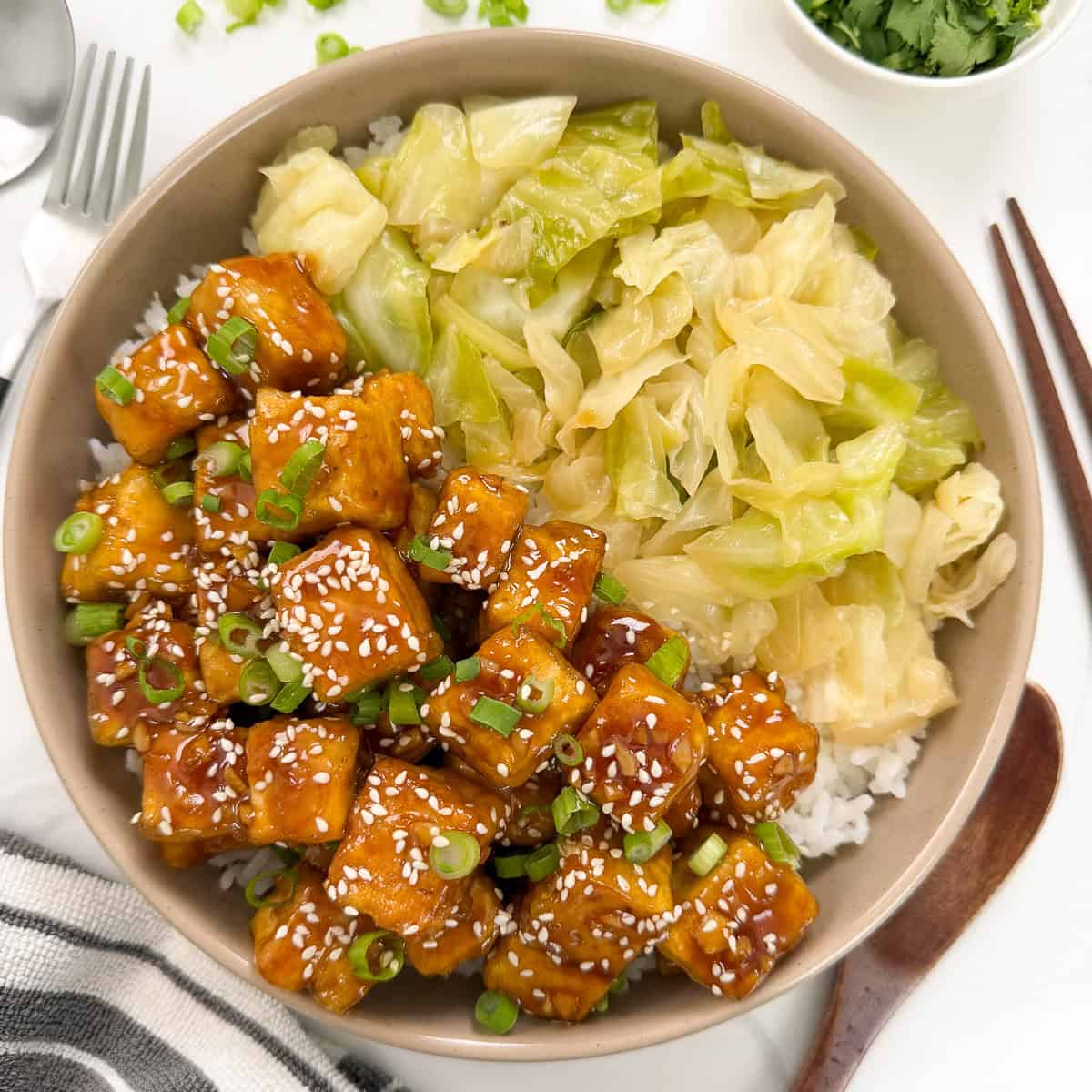 A bowl of rice topped with Honey Butter Crispy Tofu cubes, glazed and sprinkled with sesame seeds and green onions, sits alongside sautéed cabbage. Chopsticks, a fork, and a napkin are nearby.