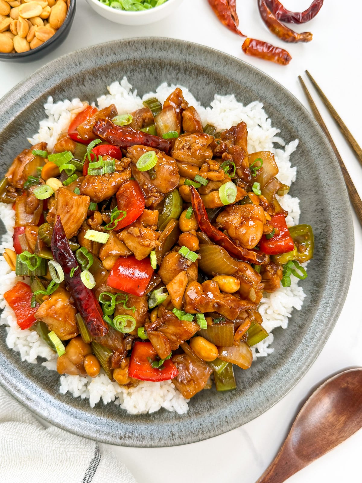 A plate of white rice topped with flavorful Kung Pao Chicken, colorful bell peppers, peanuts, dried red chilies, and chopped green onions. Bowls of peanuts and green onions are in the background. Chopsticks and a wooden spoon are beside the plate.