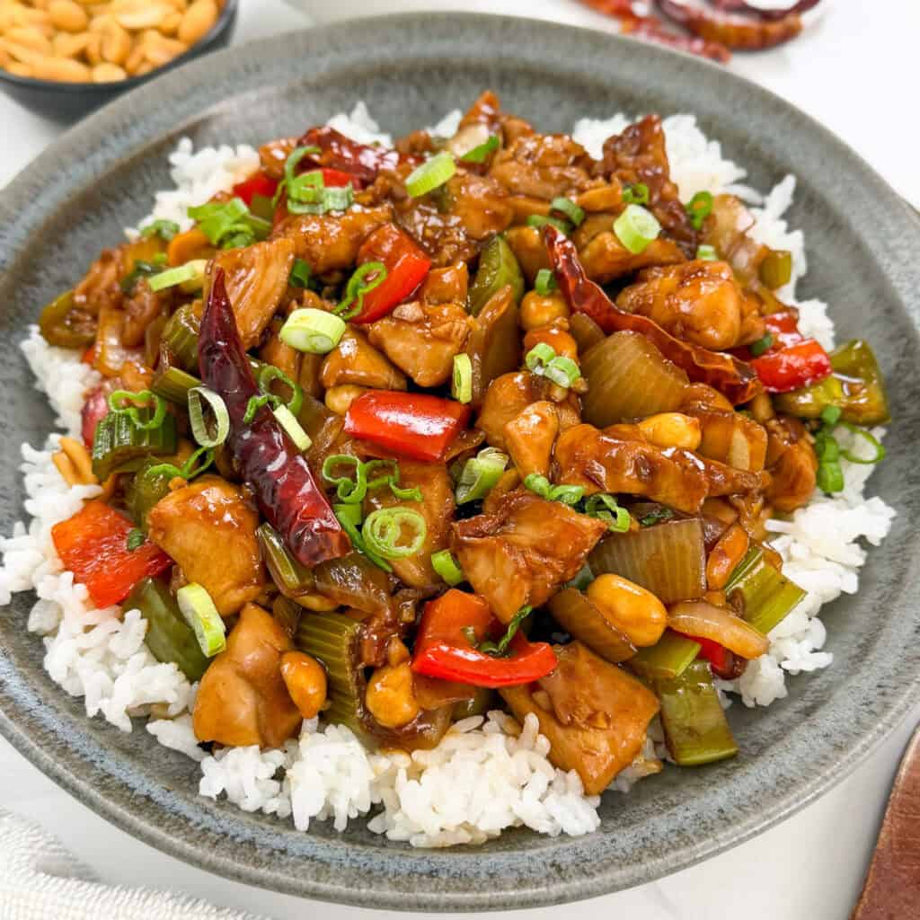 A bowl of white rice topped with classic Kung Pao Chicken, featuring diced chicken, red bell peppers, celery, peanuts, dried red chilies, and garnished with chopped green onions.