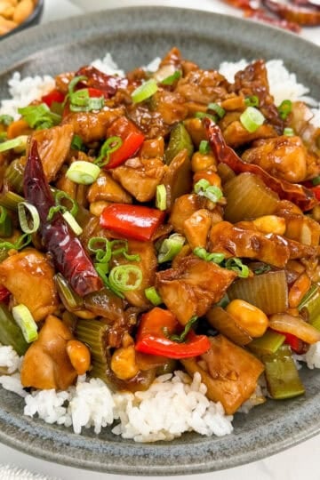A bowl of white rice topped with classic Kung Pao Chicken, featuring diced chicken, red bell peppers, celery, peanuts, dried red chilies, and garnished with chopped green onions.