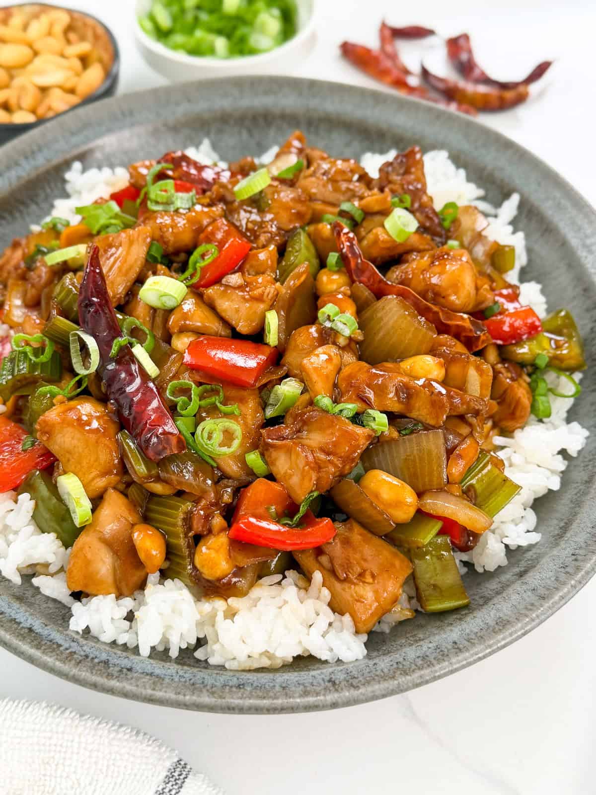 A plate of flavorful Kung Pao Chicken with red peppers, celery, peanuts, dried chilies, and green onions is served over white rice. Small bowls of peanuts and chopped green onions complete this classic Kung Pao Chicken presentation in the background.