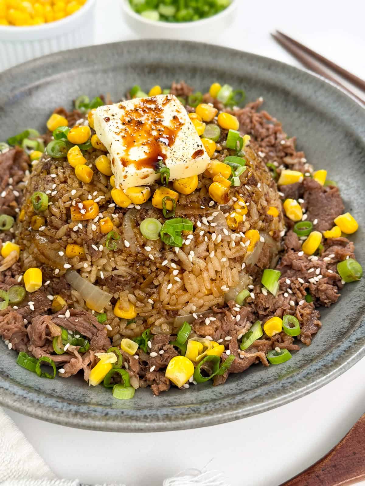 A gray plate with Rice Cooker Beef Pepper Rice (Pepper Lunch Inspired), featuring beef fried rice topped with corn, green onions, sesame seeds, and a pat of melting butter drizzled with sauce. Small bowls of green onions and corn are in the background.