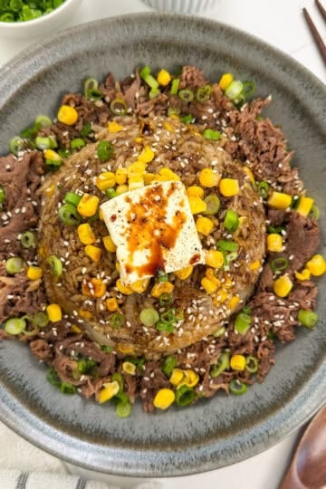A gray bowl of Rice Cooker Beef Pepper Rice (Pepper Lunch Inspired) fried rice is topped with corn, scallions, sesame seeds, a pat of butter, and soy sauce, with chopsticks and a spoon beside it.