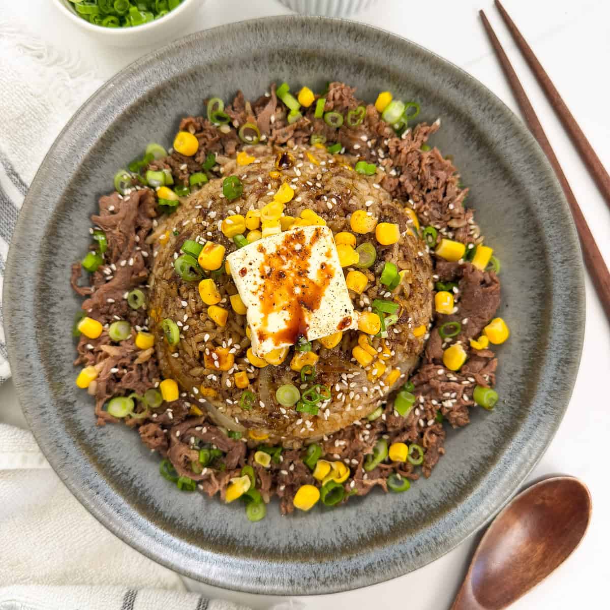 A gray bowl of Rice Cooker Beef Pepper Rice (Pepper Lunch Inspired) fried rice is topped with corn, scallions, sesame seeds, a pat of butter, and soy sauce, with chopsticks and a spoon beside it.
