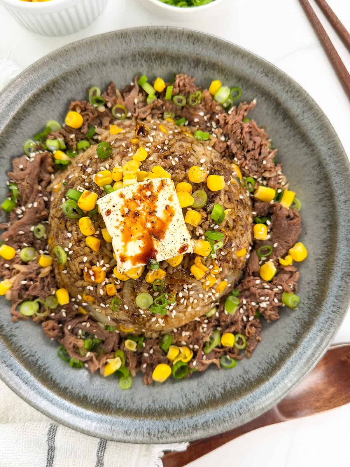 Enjoy this Rice Cooker Beef Pepper Rice (Pepper Lunch Inspired): a bowl of rice topped with corn, a pat of butter, sliced green onions, sesame seeds, and thinly sliced beef, all drizzled with savory sauce.