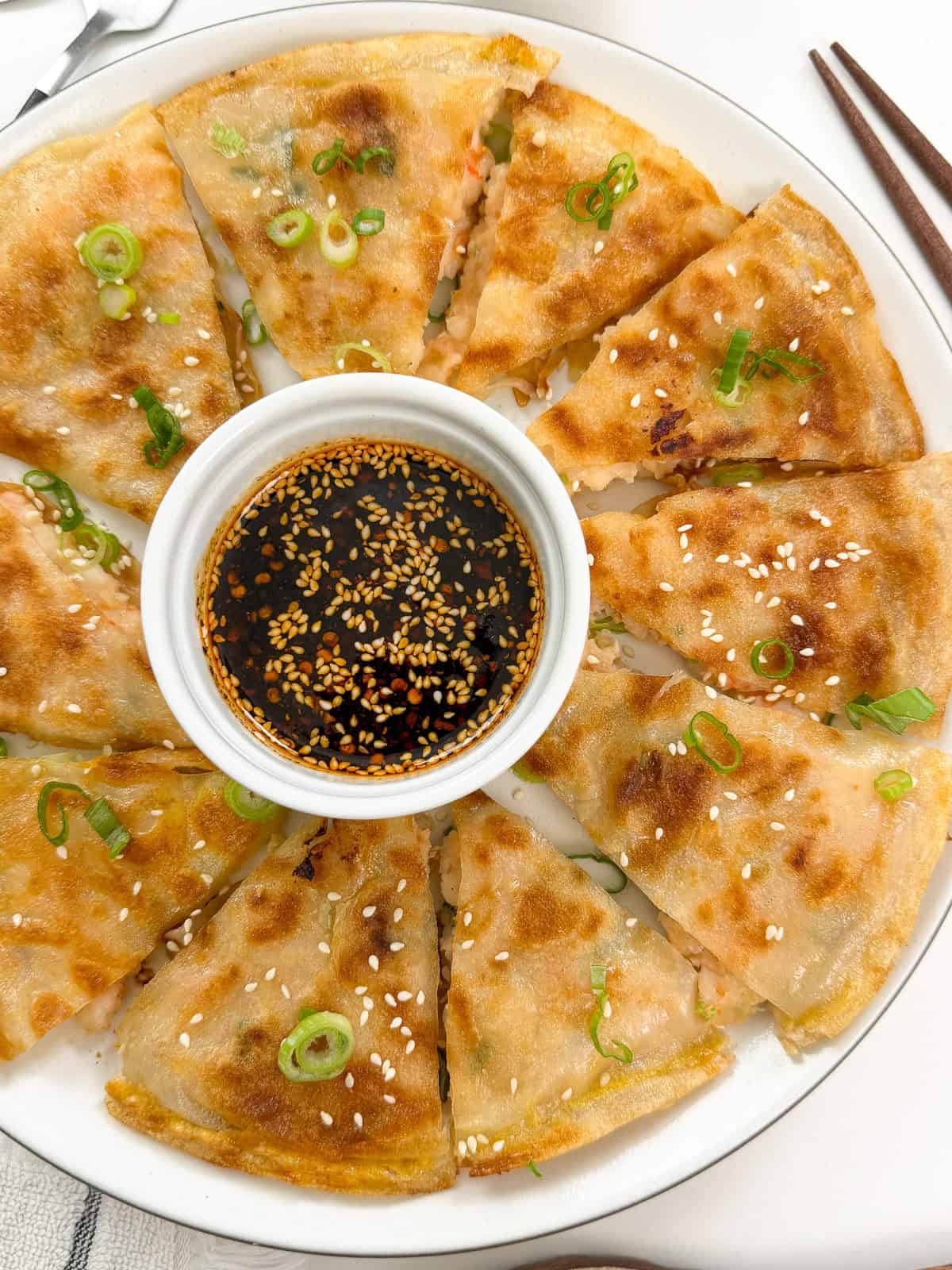 A plate of golden, crispy Rice Paper Shrimp Pancakes cut into wedges, garnished with green onions and sesame seeds, surrounds a small bowl of soy dipping sauce topped with sesame seeds. Chopsticks rest on the plate’s edge.