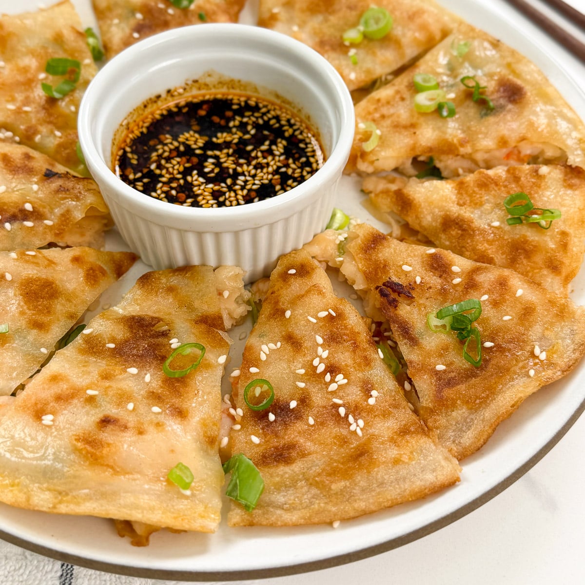 A plate of golden-brown, sliced Rice Paper Shrimp Pancakes arranged around a small dish of soy dipping sauce, garnished with sesame seeds and chopped green onions.