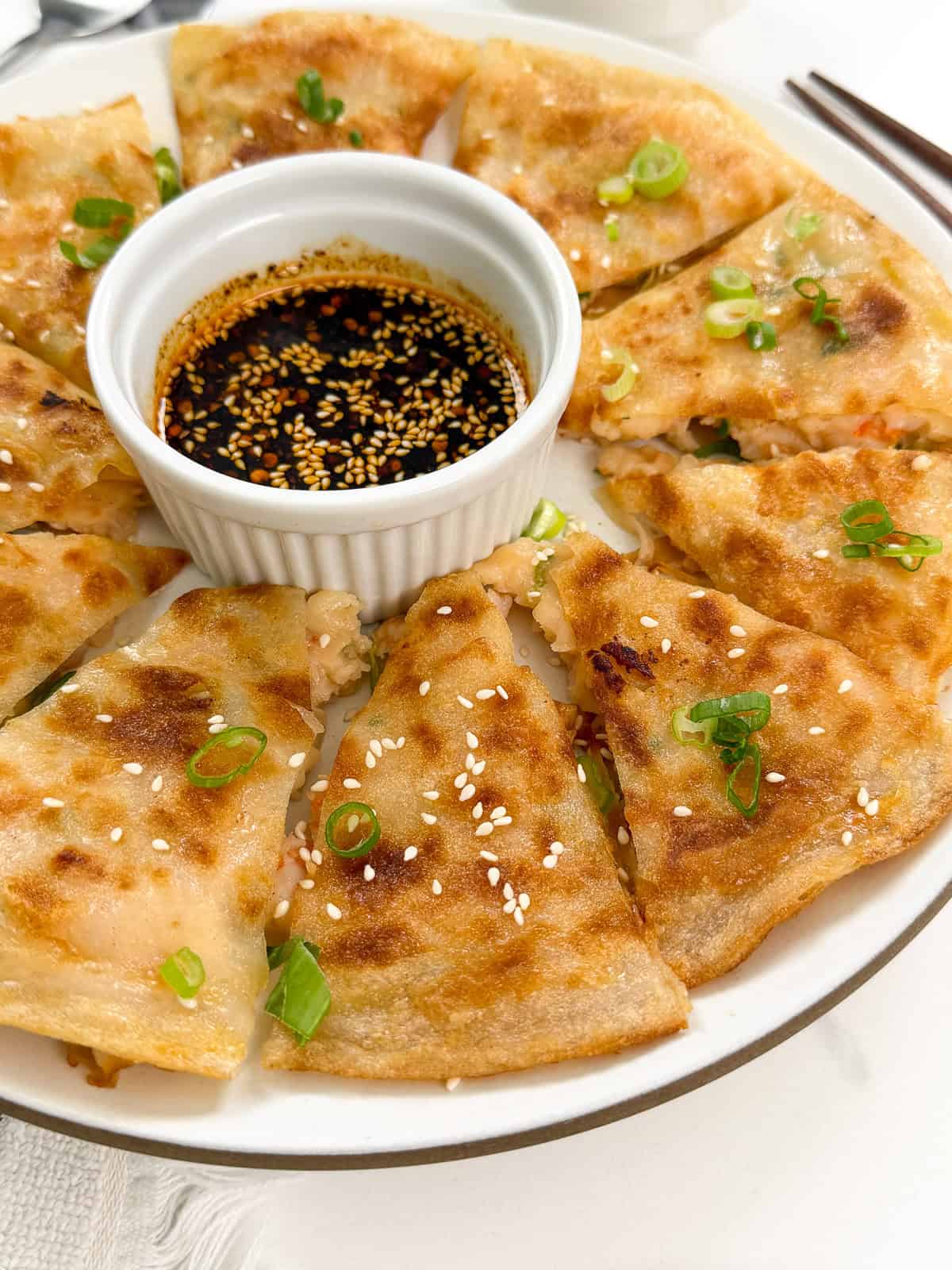 A plate of golden, crispy Rice Paper Shrimp Pancakes, sliced and garnished with green onions and sesame seeds, arranged around a small dish of dark dipping sauce.