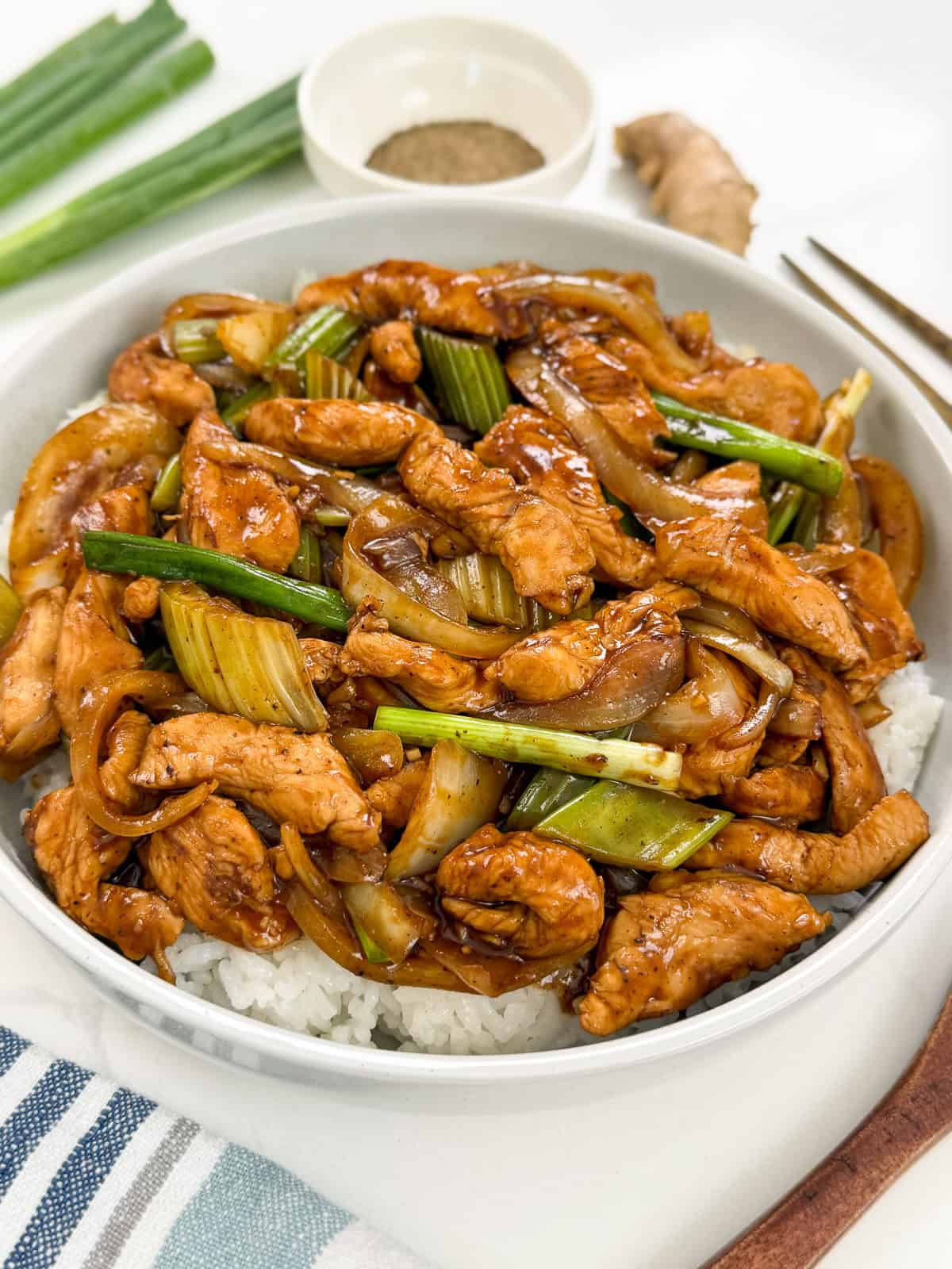 A bowl of white rice topped with Sizzling Black Pepper Chicken, featuring stir-fried chicken, sliced onions, celery, and green onions in a savory brown sauce. A dish of pepper, ginger root, and fresh green onions are in the background.