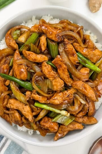 A bowl of Sizzling Black Pepper Chicken with green onions and onions in a dark sauce, served over white rice. Chopsticks and a wooden spoon are placed beside the bowl, with fresh green onions visible in the corner.