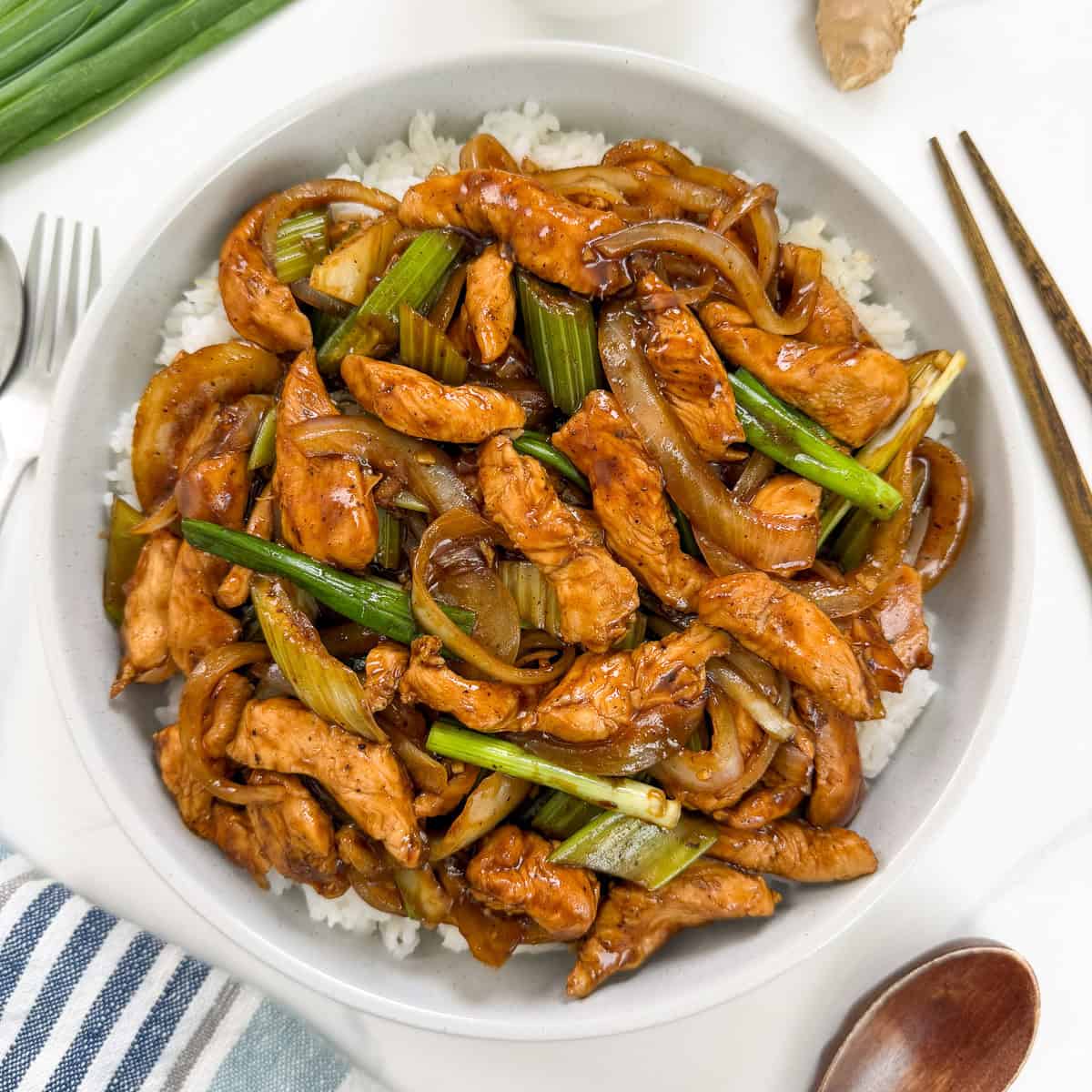 A bowl of Sizzling Black Pepper Chicken with green onions and onions in a dark sauce, served over white rice. Chopsticks and a wooden spoon are placed beside the bowl, with fresh green onions visible in the corner.