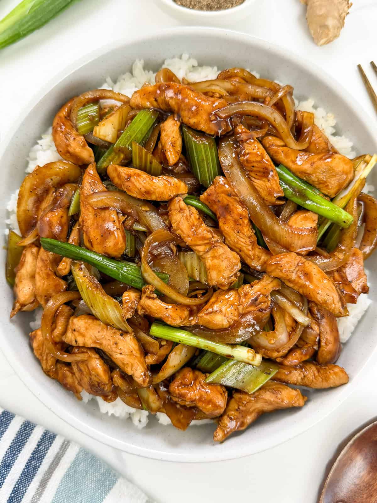 A bowl of Sizzling Black Pepper Chicken with sliced onions and green onions in a savory brown sauce, served over white rice. The dish is arranged neatly and looks flavorful and freshly cooked.