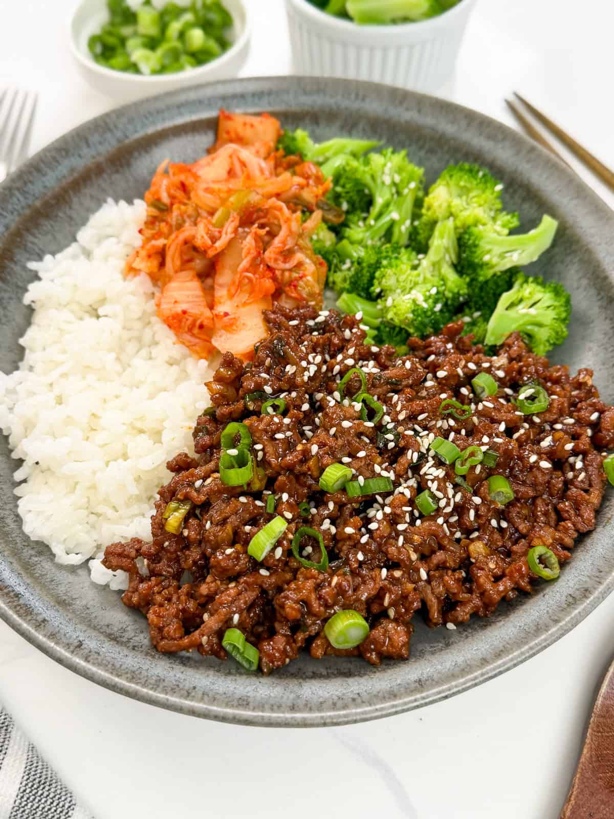 A plate with white rice, spicy Sticky Gochujang Ground Beef Bowls topped with sesame seeds and green onions, steamed broccoli, and kimchi. Small bowls of green onions and cucumbers are in the background.