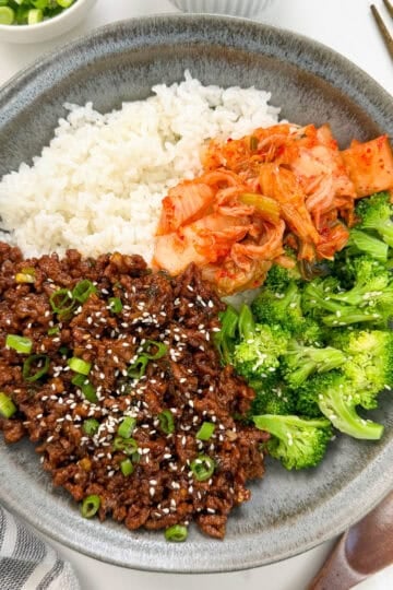 A gray plate with white rice, kimchi, steamed broccoli, and Sticky Gochujang Ground Beef Bowls topped with green onions and sesame seeds. Chopsticks, a wooden spoon, and a striped napkin are beside the plate.