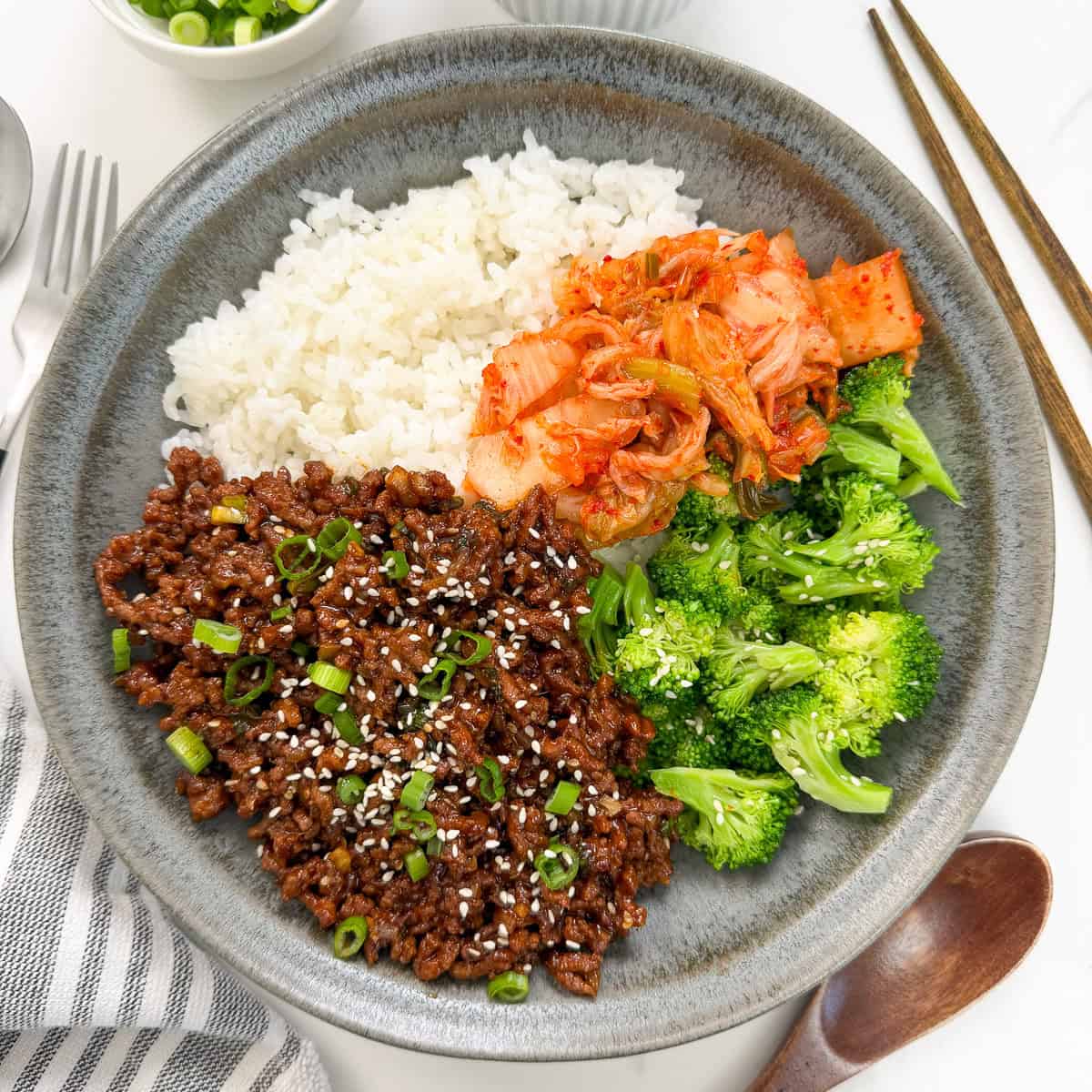 A gray plate with white rice, kimchi, steamed broccoli, and Sticky Gochujang Ground Beef Bowls topped with green onions and sesame seeds. Chopsticks, a wooden spoon, and a striped napkin are beside the plate.
